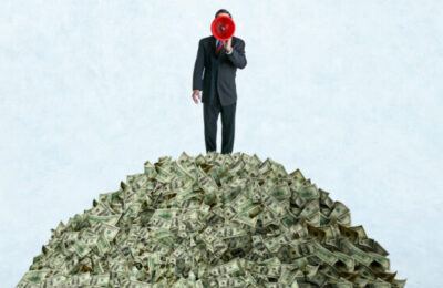 A businessman shouts through a red megaphone as he stands on top of a pile of money.